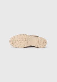Chaussures beige avec une semelle en caoutchouc texturé présentant un motif de bande de roulement. La semelle est épaisse et conçue pour offrir adhérence et durabilité.