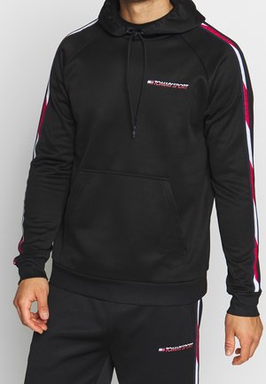 Mand iført sort Tommy Sport træningsdragt med røde og hvide striber på ærmer og bukser, stående mod en ensfarvet baggrund.