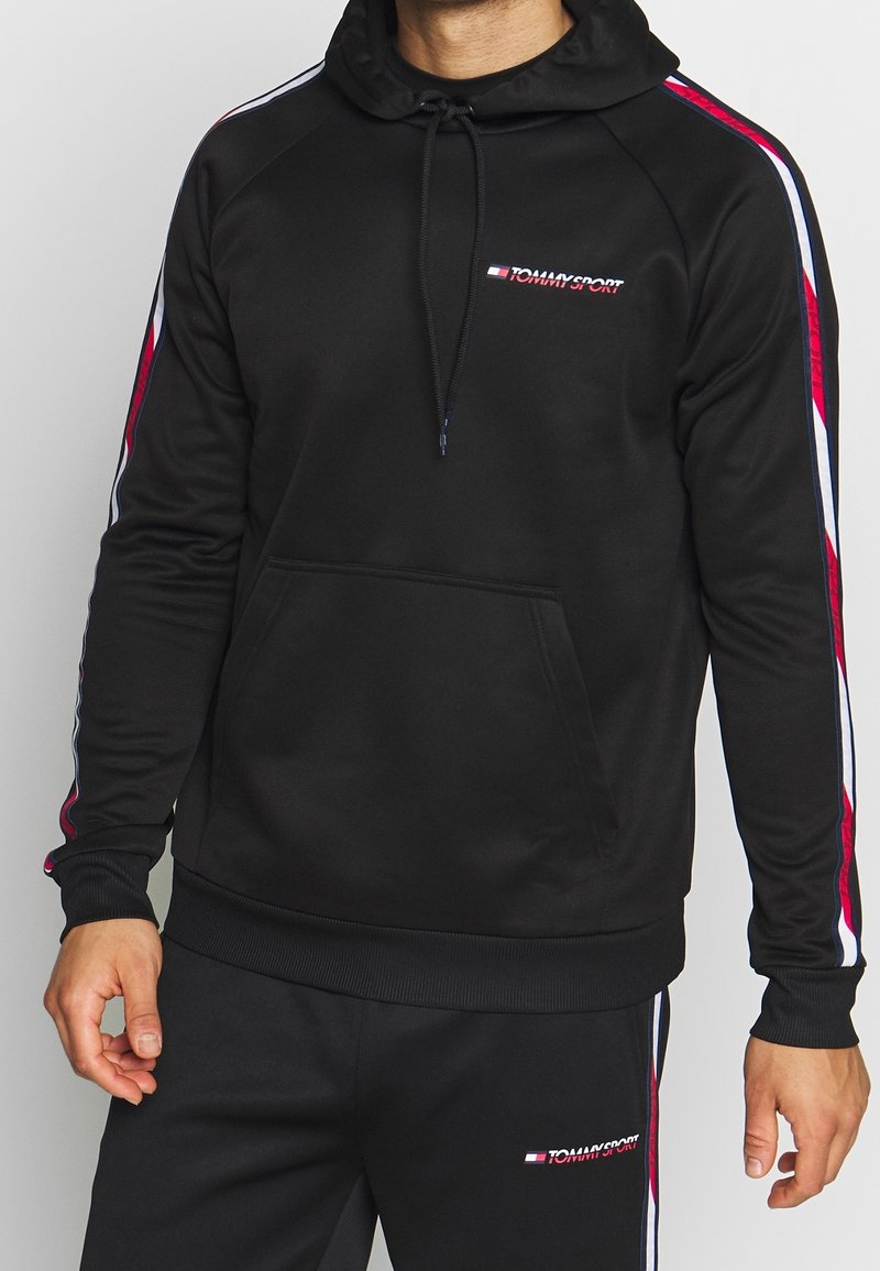 Homme portant un survêtement Tommy Sport noir avec des bandes rouges et blanches sur les manches et le pantalon, debout devant un fond uni.