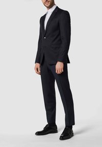 Marineblauer Anzug mit taillierter Passform, Reverskragen, einreihigem Knopfverschluss und passenden marineblauen Hosen. Schwarze Lederschuhe vervollständigen das Outfit.