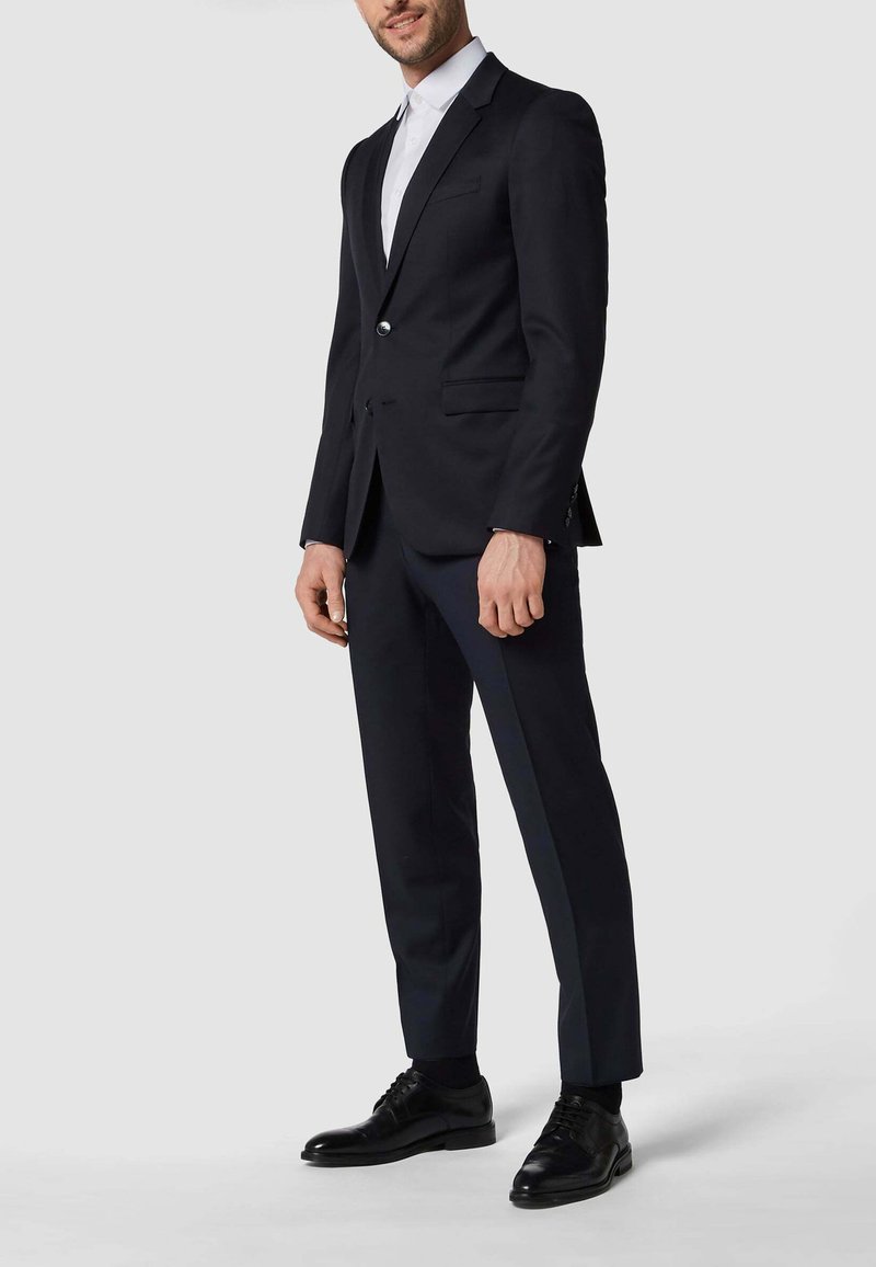 Marineblauer Anzug mit taillierter Passform, Reverskragen, einreihigem Knopfverschluss und passenden marineblauen Hosen. Schwarze Lederschuhe vervollständigen das Outfit.