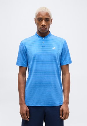 Άνδρας με κοντά ξανθά μαλλιά, που φοράει μπλε ριγέ πόλο μπλουζάκι Adidas και σκουρόχρωμο παντελόνι, στέκεται μπροστά σε ένα απλό ανοιχτό φόντο.