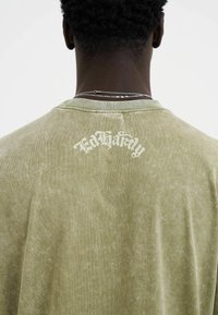 Zelené, texturované tričko s krátkými rukávy, které má jemně vybledlý vzhled a bílé logo "Ed Hardy" na zadní straně.