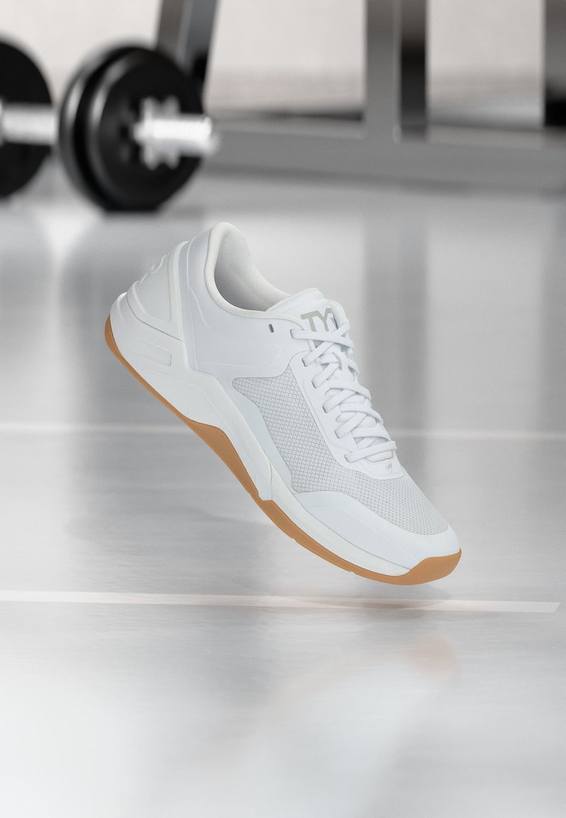 Λευκό αθλητικό παπούτσι με καουτσούκ σόλα που αιωρείται σε εσωτερικό χώρο, ο εξοπλισμός γυμναστηρίου είναι θαμπός στο φόντο.