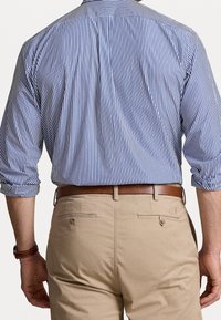 Man som bär en uppvikt blå och vit randig skjorta instoppad i beige byxor med ett brunt läderbälte, sedd från baksidan.