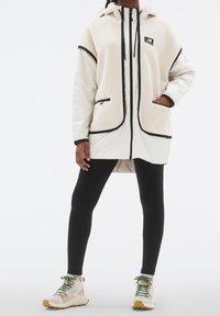 Lätt, beige fleecejacka med svarta detaljer, zip-fickor och huva, kombinerad med svarta leggings och krämfärgade sneakers.