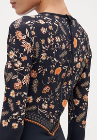 Haut à manches longues bleu marine avec des motifs floraux orange et crème. Doté d'une fermeture éclair à l'arrière et de détails texturés au niveau de l'ourlet à la taille.