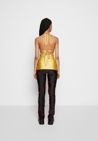 Top halter amarelo de cetim com atilhos nas costas, combinado com calças pretas com padrões cruzados vermelhos e sapatos de plataforma.