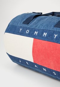 Denimová cestovná taška s námorníckymi rukoväťami. Obsahuje biely a červený vzor s textom "TOMMY" a "JEANS" v bielej farbe. Guľatý tvar, štruktúrovaná látka.