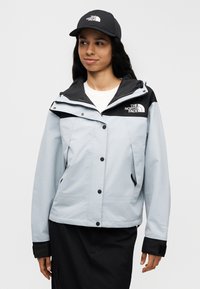 Ελαφρύ γαλάζιο μπουφάν με μαύρη κουκούλα και διακοσμητικά στοιχεία, με κουμπιά και τσέπες, συνδυασμένο με μαύρο καπέλο που φέρει το λογότυπο της The North Face.