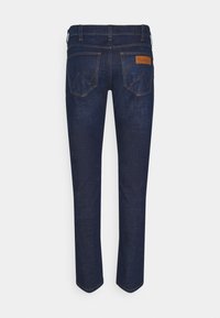 Tummansinisiä farkkuja, suora leikkaus, jossa on kaksi takataskua ja vaaleanruskea nahkalappu, jossa on brändin nimi. Eroava ompelu näkyvissä.