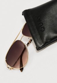 Solbriller med guldramme og gradientbrune linser med skildpaddemønstrede detaljer på stængerne, ved siden af et sort læderetui prydet med "QUAY."