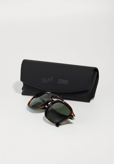 Γυαλιά ηλίου με καφέ καβούκι και σκούρους φακούς, συνοδευόμενα από μια μαύρη δερμάτινη θήκη με ανάγλυφο λογότυπο.