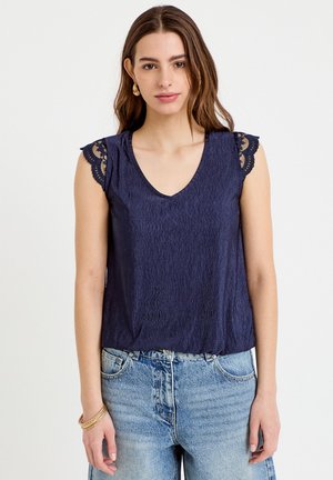 Kobieta o długich brązowych włosach, ubrana w granatowy, teksturowany, bezrękawnik z koronkowymi krótkimi rękawkami i jasnoniebieskie, wysokowysokie jeansy, stojąca na białym tle.