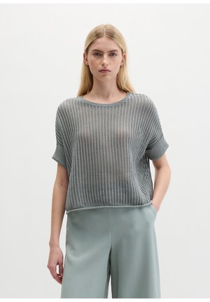 Young woman with straight blonde hair wearing a loose knit short-sleeve gray sweater and light blue wide-leg pants, standing with hand in pocket.