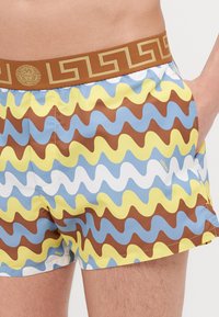 Kupaće hlače s raznobojnim valovitim uzorkom u plavoj, žutoj, smeđoj i bijeloj boji, s smešnim pojasom ukrašenim zlatnim logotipom.
