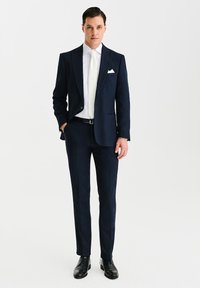 Granatowy wełniany garnitur o dopasowanym kroju, z jedno-guzikową marynarką, klapami w kształcie notch i białą koszulą. Uzupełniony białym krawatem i poszetką.