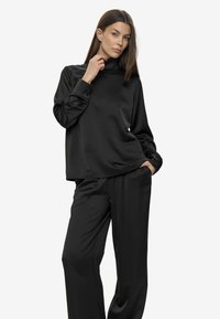 Femme aux longs cheveux bruns, portant un haut à col roulé en satin noir et un pantalon large, posant avec une main près de son cou et l'autre dans sa poche.
