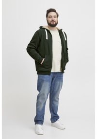 Sudadera con cremallera de color verde oscuro y forro beige, con dos bolsillos, cordones ajustables, combinada con jeans de color azul claro y zapatillas blancas.