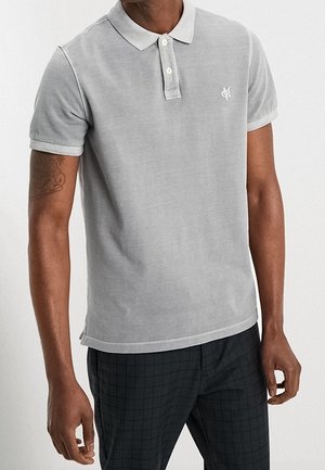 Polo grigia con colletto, maniche corte e logo bianco sul lato sinistro. Indossata con pantaloni scuri a quadri, con orlo dritto.