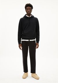 Zwarte hoodie met kangoeroezak, witte binnenkraag, zwarte corduroy broek en beige suède pantoffels. Model staat tegen een neutrale achtergrond.