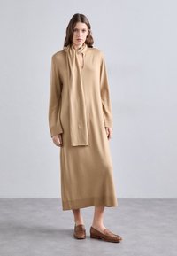 Robe beige en maille avec manches longues, encolure fendue et détail d'écharpe assorti. Portée avec des mocassins marron sur un fond gris clair.