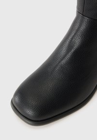 Bota de tornozelo em couro preto com um acabamento texturizado, bico quadrado arredondado e design minimalista. Sola fina com detalhes sutis nas bordas.