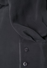 Svart blus med ett följsamt tyg och en mjuk textur, med knäppte ärmslut och mjukt samlade ärmar.