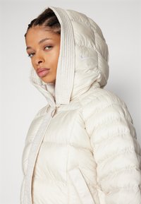 Gefütterte weiße Kapuzenjacke mit Steppdesign, ausgestattet mit einem Frontreißverschluss und Seitentaschen, akzentuiert durch ein kleines Nike-Logo auf der Kapuze.