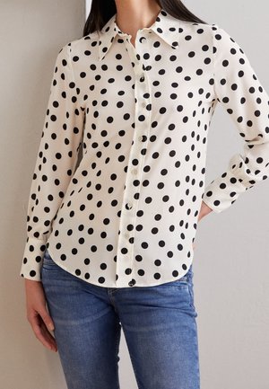 Vrouw draagt een wit, lang mouwtjes, knoopshirt met zwarte stippen en blauwe jeans, staat met een hand op haar heup.