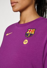 Ljubičasta košulja dugih rukava s rebrastom teksturom, s žutim Nike logotipom, vezanom grbom FC Barcelone i žutim Spotify simbolom.
