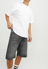 Henkilö, jolla on päällään yksinkertainen valkoinen T-paita, mustat kulutettu denim-shortsit ja valkoiset sukat mustilla raidoilla, seisoo yksinkertaista taustaa vasten.