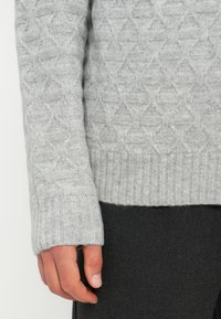Pull en tricot gris clair avec un motif géométrique texturé et des poignets côtelés, associé à un tissu gris foncé visible en bas.