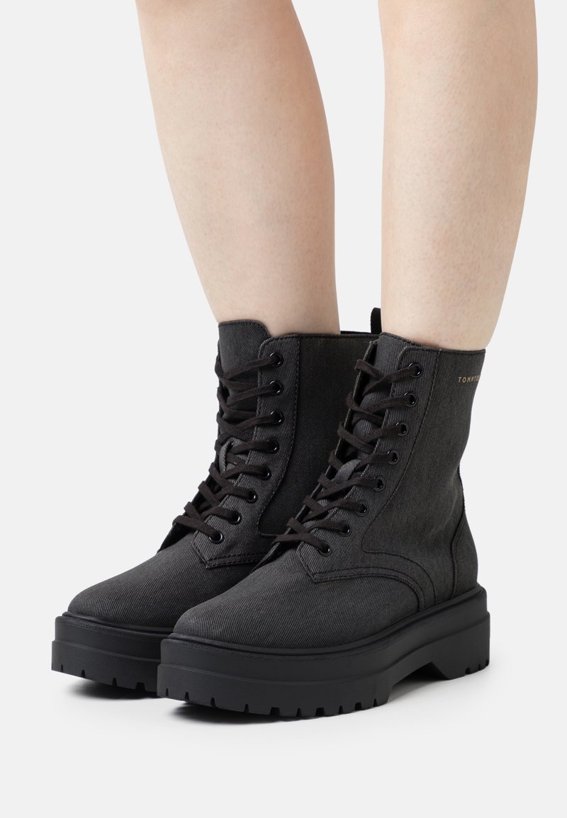 Bottes de combat noires à lacets avec semelles épaisses portées sur des jambes nues, photographiées sur un fond clair uni.