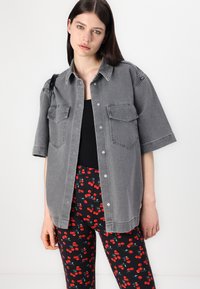 Grå denimskjorte med korte ermer, knappestolpe foran og to brystlommer, kombinert med svarte bukser med et rødt kirsebærmønster.