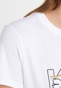 Valkoinen puuvillapaita, jossa on musta graafinen design kullan kimaltavalla korostuksella. Lyhyet hihat ja pehmeä tekstuuri. Rentoa istuvuutta.