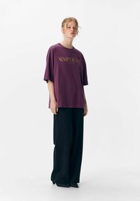 T-shirt violet surdimensionné avec le texte "Simplicité" en or. Assorti à un pantalon large noir, sur un fond clair et uni.