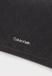 Accessoire en cuir noir texturé Calvin Klein avec motif géométrique embossé et lettrage argenté du logo de la marque.