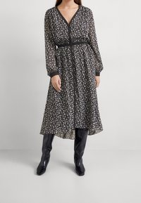 Musta kukkakuvioinen midi mekko, jossa on V-pääntie, pitkät puffihihat ja vyötäröltä kinnautuva muoto. Kirkas kangas, jossa on pieniä valkoisia kukkakuviota. Yhdistetty mustiin saappaisiin.