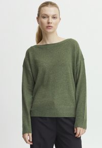 Grüner Strickpullover mit weitem Bootsausschnitt, fallenden Schultern und gerippten Bündchen, aus weichem, strukturiertem Stoff. Kombiniert mit dunklen Hosen.