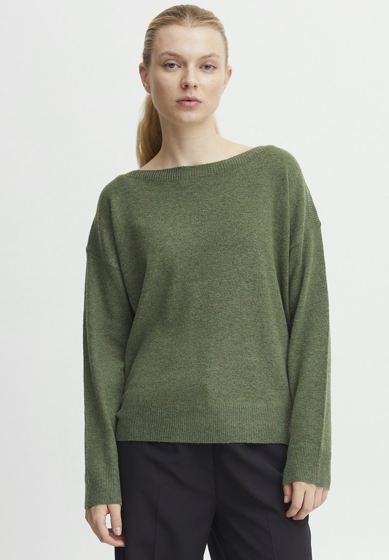 Grüner Strickpullover mit weitem Bootsausschnitt, fallenden Schultern und gerippten Bündchen, aus weichem, strukturiertem Stoff. Kombiniert mit dunklen Hosen.