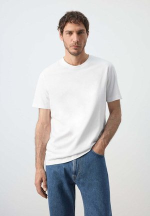 Man draagt een effen wit T-shirt met korte mouwen en blauwe spijkerbroek, staand tegen een lichtgrijze achtergrond met één hand in de zak.