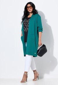 Cardigan a maglia color teal con due tasche sul fronte, indossato sopra una maglietta nera con un motivo metallizzato, abbinato a pantaloni bianchi a tre quarti e una borsa nera.