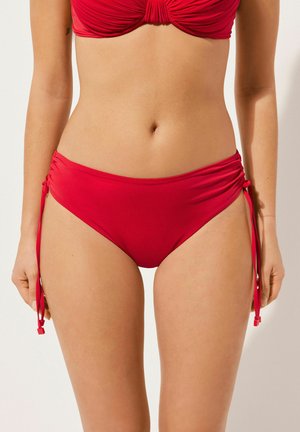 Bas de bikini rouge en tissu lisse avec des liens sur les côtés et des détails froncés. Présente une jambe haute pour un ajustement flatteur.