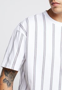 White short-sleeve t-shirt with vertical stripes in navy, red, and black. Features a crew neckline and subtle texture.
