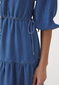 Robe bleue boutonnée devant avec jupe à volants froncée, taille élastique resserrée par un lien latéral et manches bouffantes mi-longues sur un bras à peau claire.