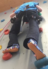 Kletterwandgurt mit bunten Kletterschuhen; die Schuhe haben Cartoon-Muster und Gummisohlen, Nylonriemen sichern den Gurt um die Taille.