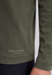 Olivgrünes Langarmshirt aus strukturiertem Stoff, mit einem subtilen Markenlogo in heller Schrift am unteren Saum.
