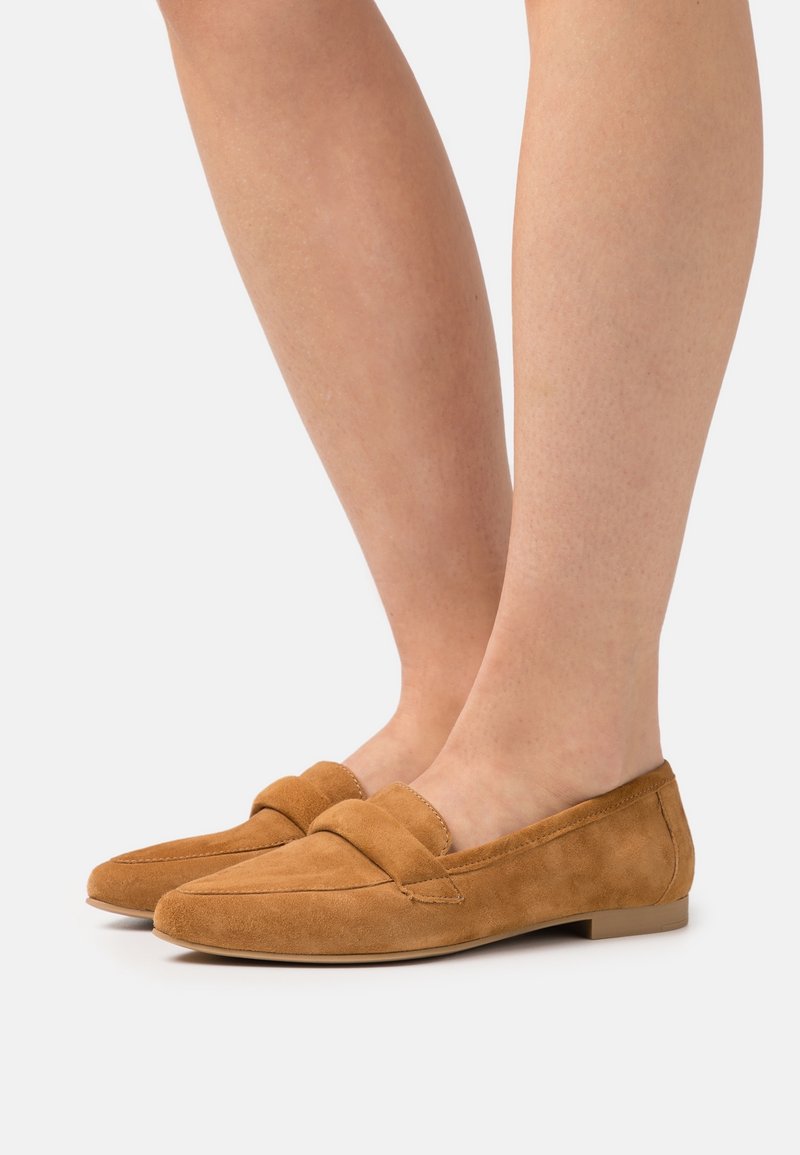 Pa paire de mocassins en daim beige portés pieds nus, montrant les vues latérale et frontale avec un petit talon sur un fond clair uni.