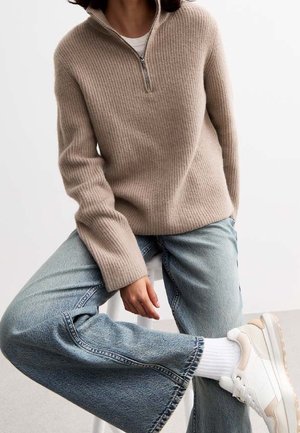 Personne portant un pull en maille beige zippé, un jean large bleu clair, des chaussettes blanches et des baskets blanches, assise avec une jambe croisée sur l'autre.